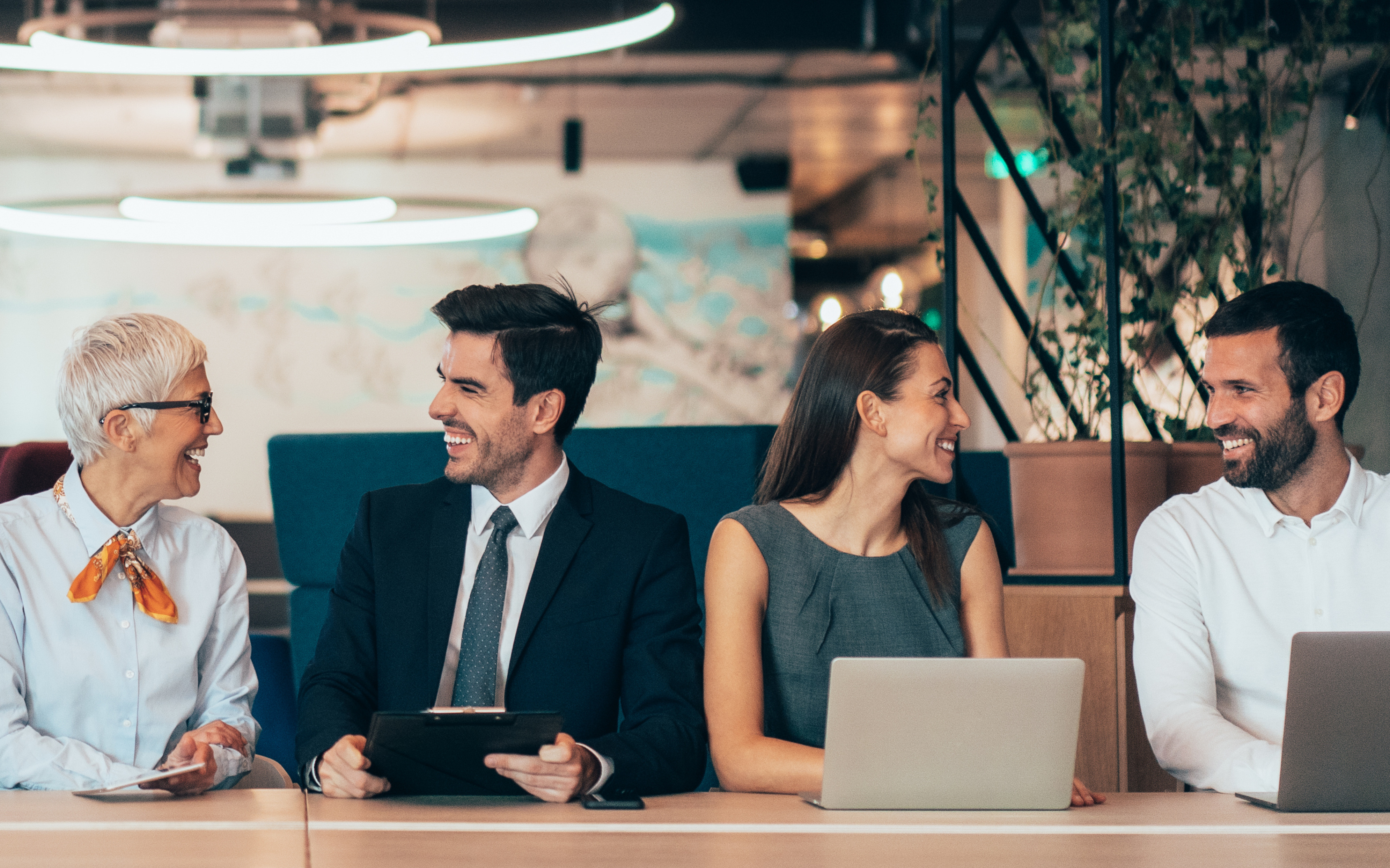 Dos pares de personas, una señora  mayor y un hombre de media edad con corbata se miran y sonríen, otra mujer de media edad se mira con otro hombre de media edad con barba y también se sonríen. Todos andan vestidos formal y están en un lugar de coworking.